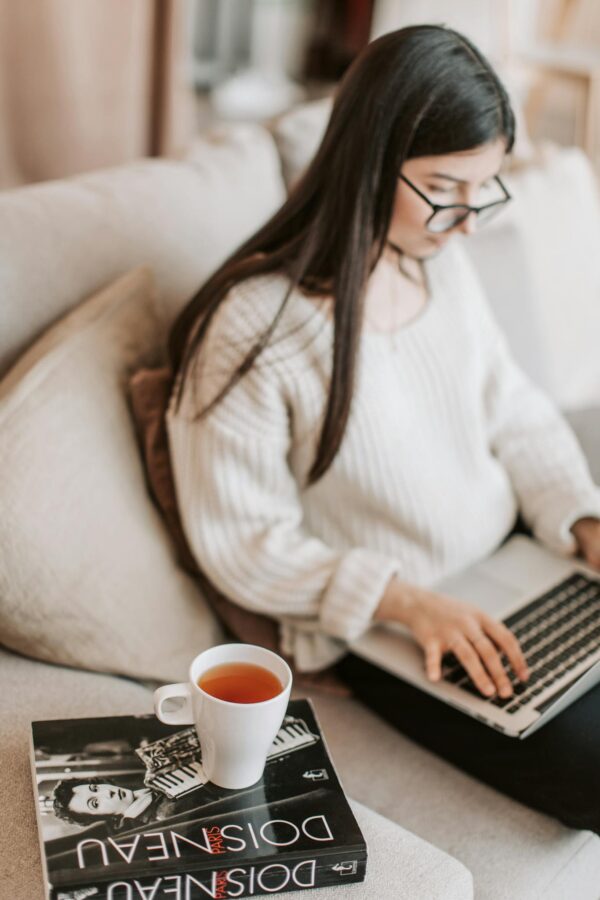 Young woman in eyeglasses using laptop on sofa with cup of tea